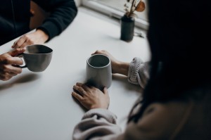 Photo of two figures sitting at a table facing each other. Their hands are on the table, curled around mugs. Their shoulders and heads are out of focus but it suggests a conversation.