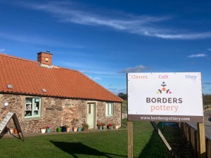 A photo of the Borders Pottery workshop, a low stone building with a red tile roof, against a blue sky.