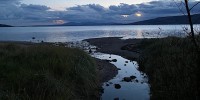 A photo of a small creek flowing into the sea. The bed of the creek is black because the sky is darkening. There are clouds on the horizon.